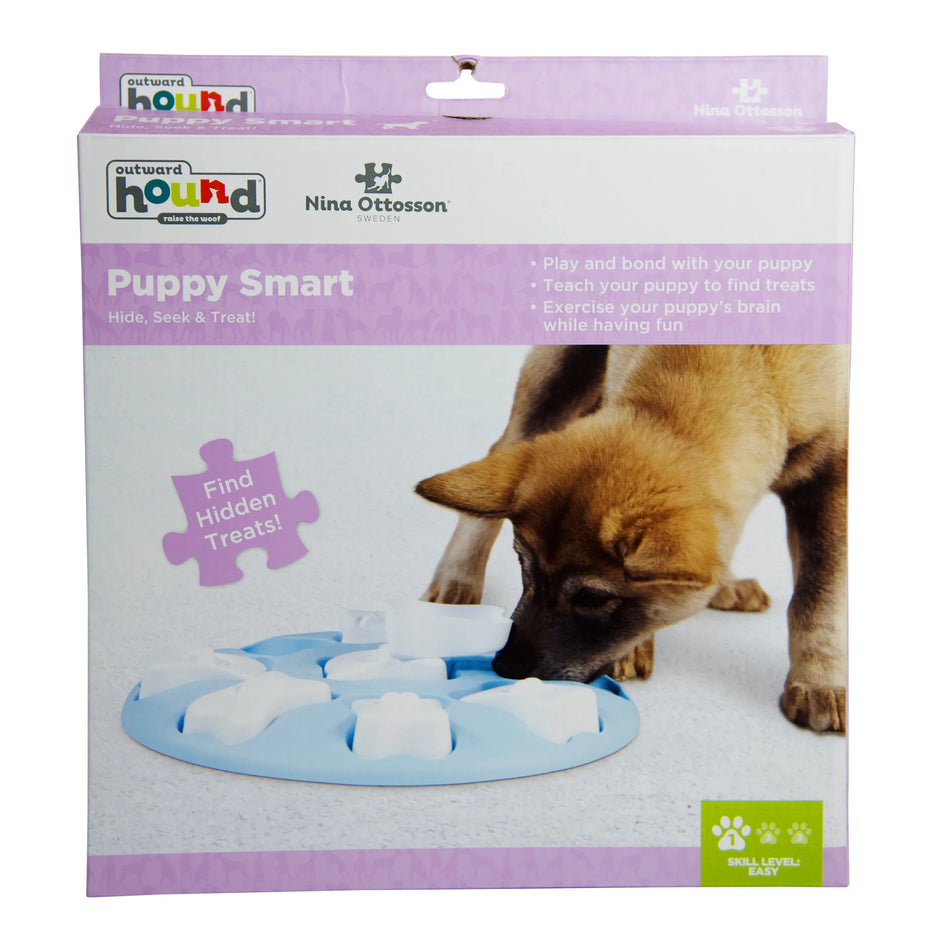 Outward Hound Nina Ottosson Puppy Smart interactive puzzle toy in blue, shown in packaging with a puppy nosing the board