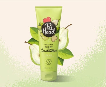 Pet Head Mucky Pup Puppy Conditioner 250ml tube in light green packaging, surrounded by illustrated pear and leaves on a pale background.