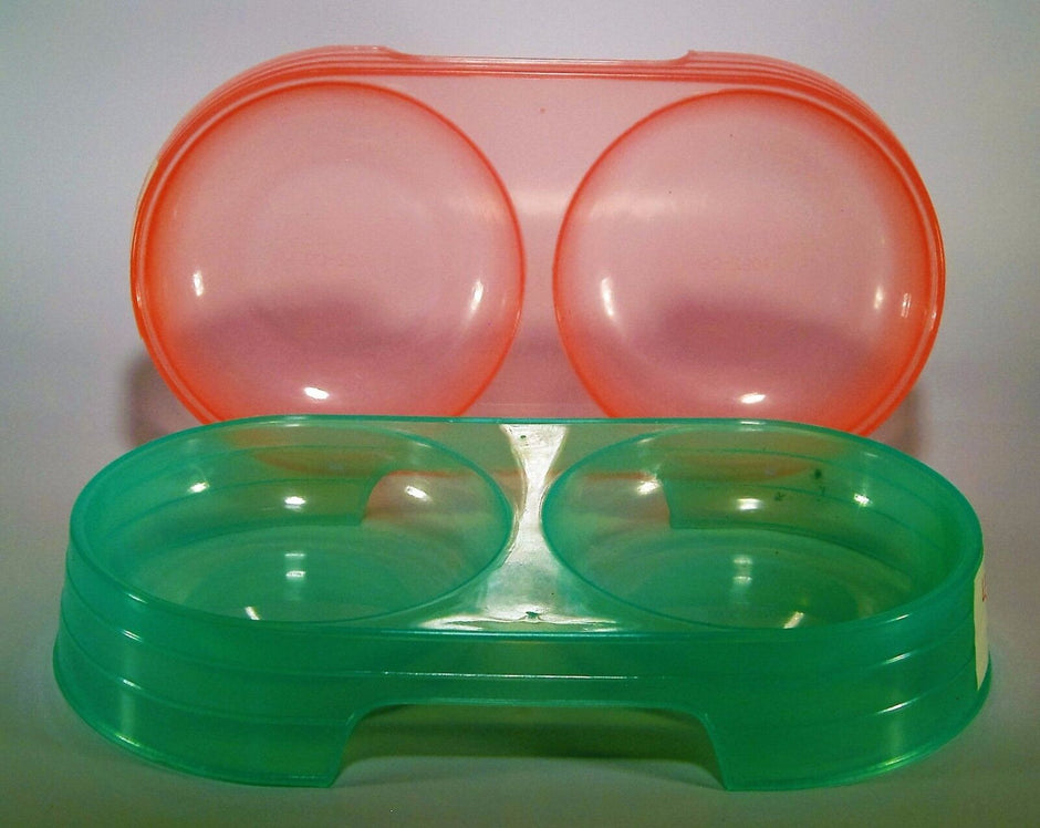 Lessmar double plastic pet bowl shown in both available colours — orange (rear) and green (front), each with two round compartments for food and water.