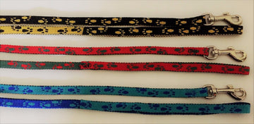 Five GoPetPlace paw print training leads laid flat, showing colour variants including black/gold, yellow/black, red/black, green/red, teal/blue, and blue, each with a silver steel swivel clip.